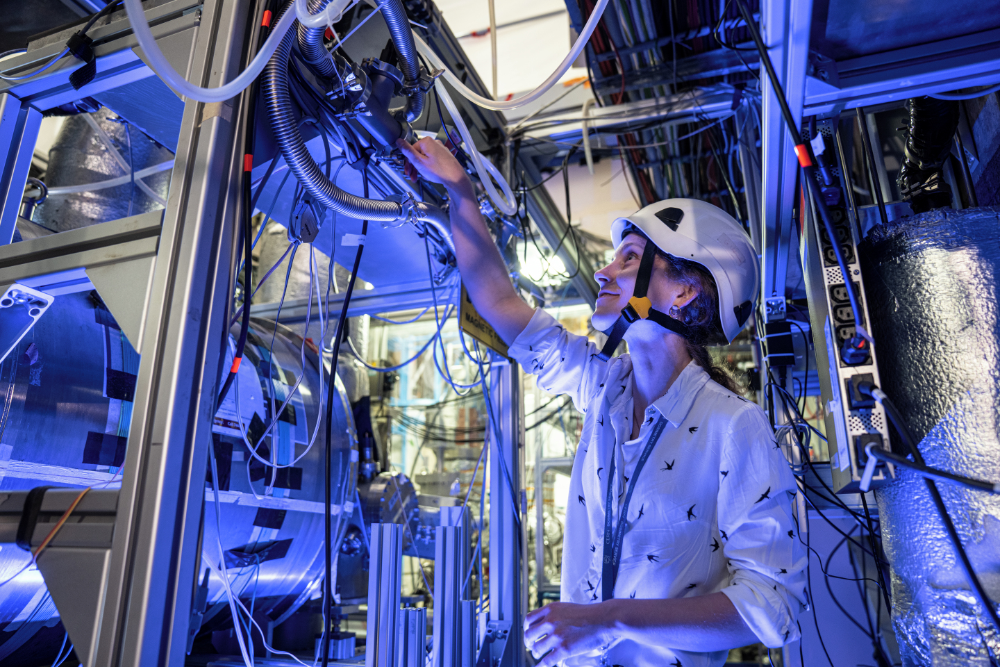 Female physicist with a helmet in front of a silver cryostat surrounded by many cables and pipes